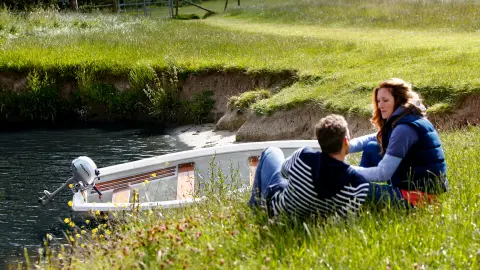 Twp people sitting on a river bank next to a boat in the water.